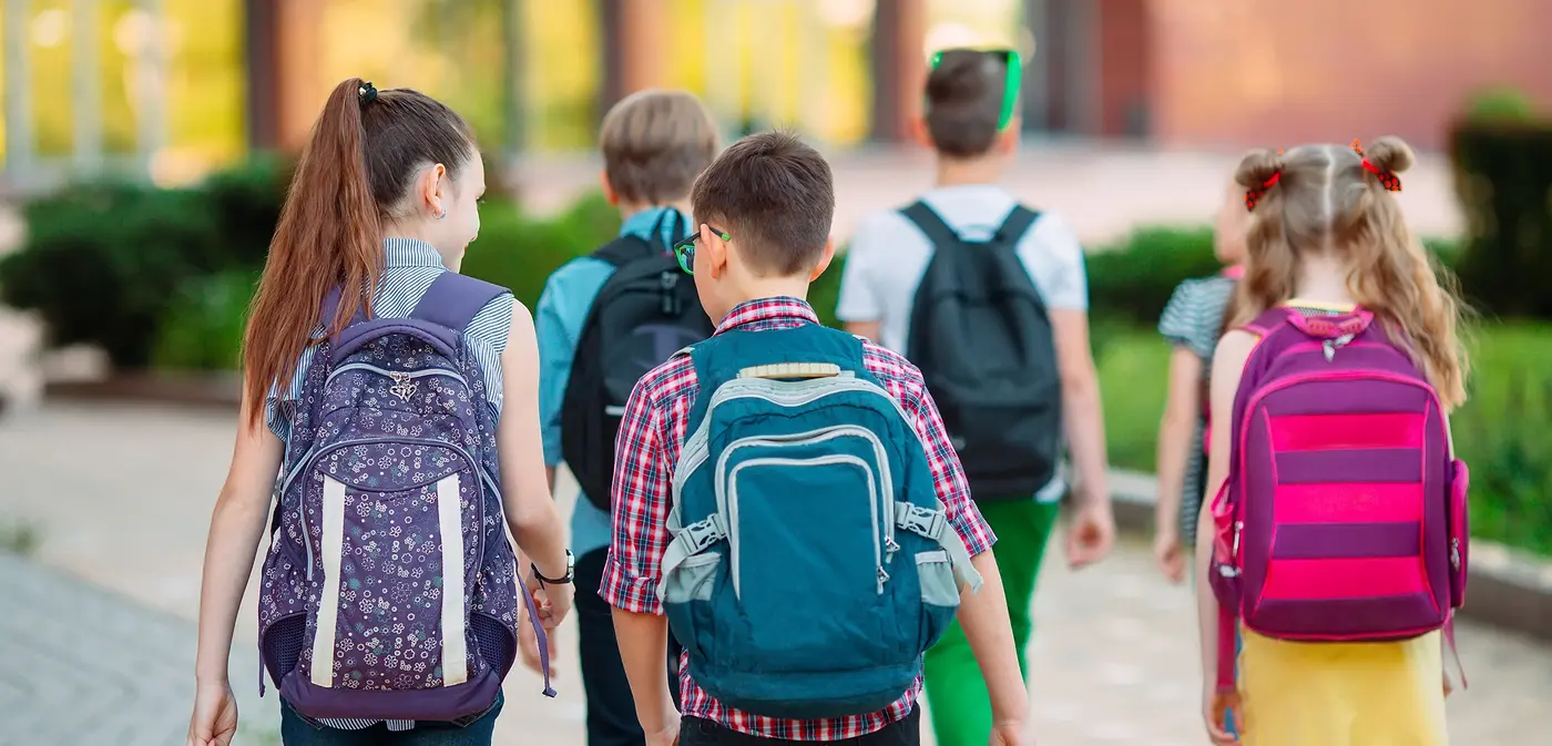 Eine Gruppe Kinder mit Rucksäcken geht zur Schule. Man sieht die Kinder von hinten.